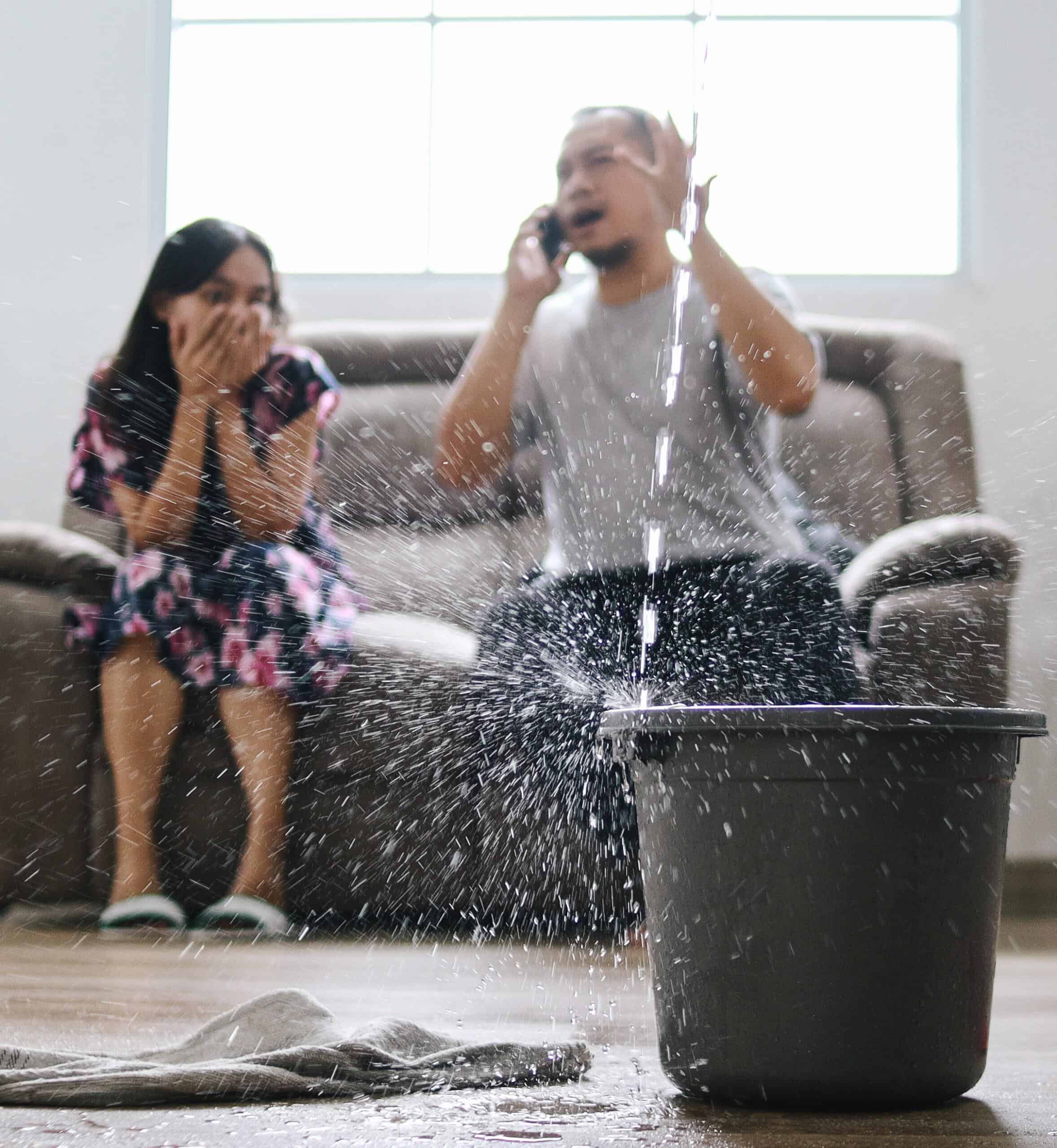 A panicking couple sitting on their living room couch panics on the phone as a roof leak fills up a water bucket.